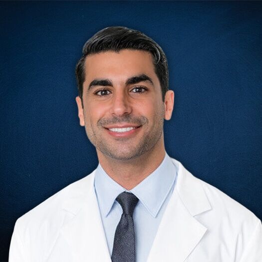 A smiling man with dark hair, wearing a white lab coat, light blue shirt, and dark tie, stands in front of a plain dark blue background.